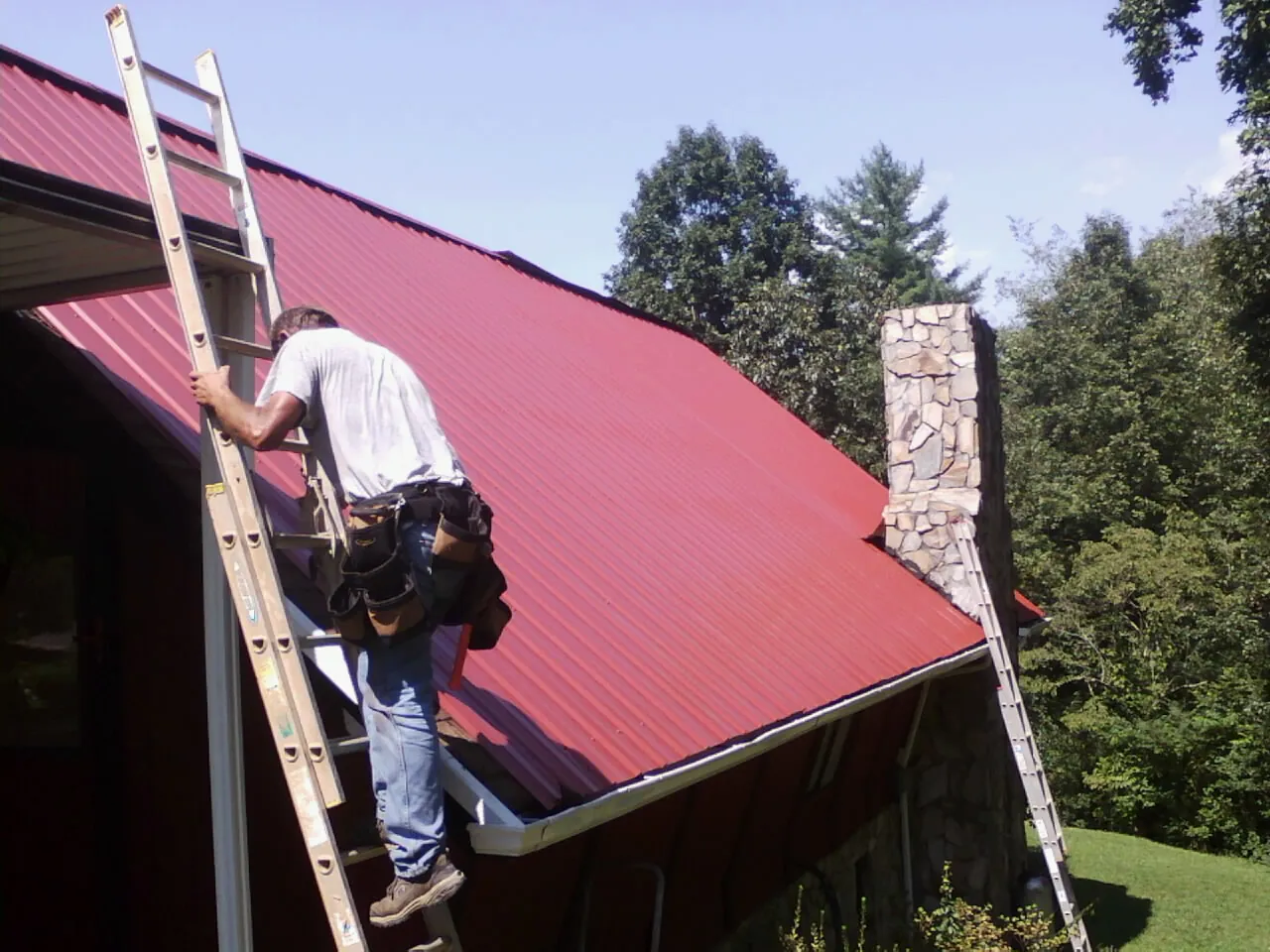 Roofer on a ladder beside a red metal roof and stone chimney — Soffit & Fascia Repair by Roofing Brothers in Gwynedd Valley